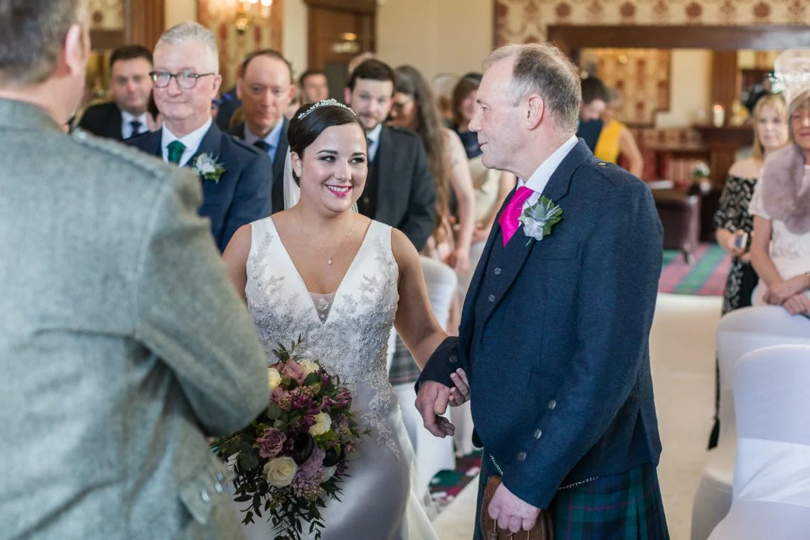A bride in a white v-neck dress smiles during her wedding ceremony as she holds hands with an older man in a kilt, surrounded by guests.