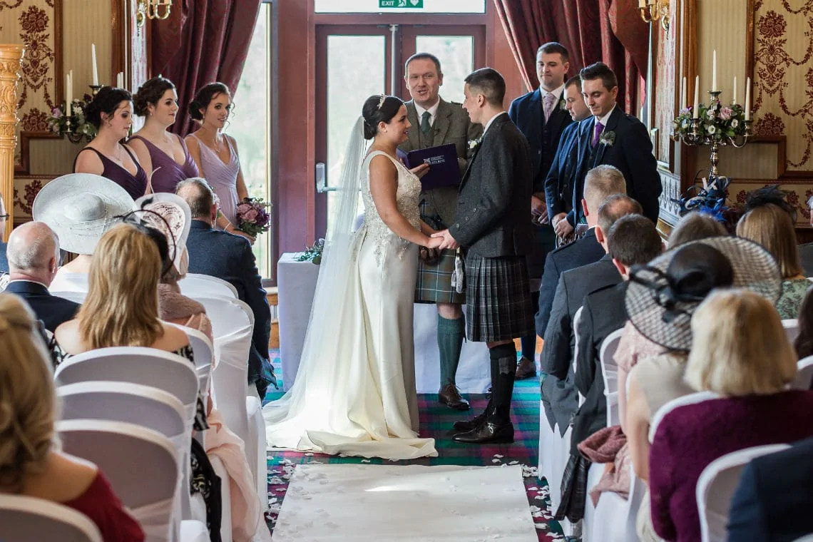 A wedding ceremony in a decorated room with guests watching as the bride in a white gown and the groom in a kilt stand facing each other.