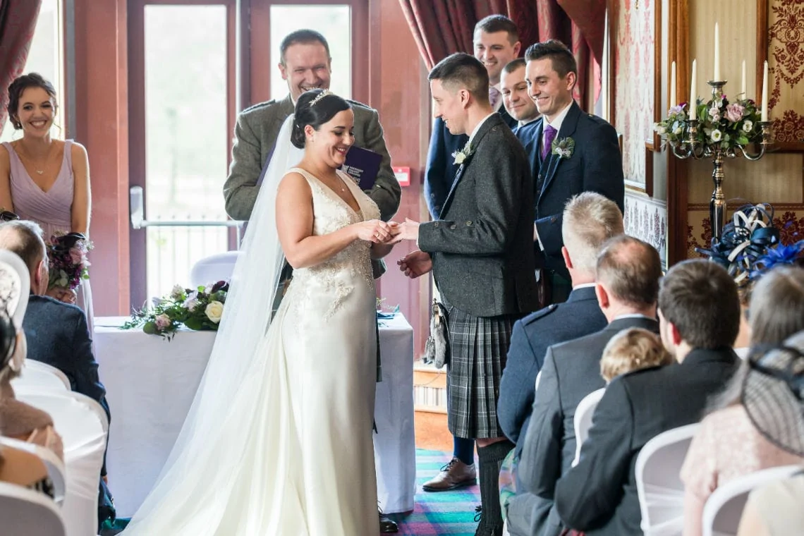 Bride and groom exchanging rings at a wedding ceremony, with guests and a smiling officiant in the background.