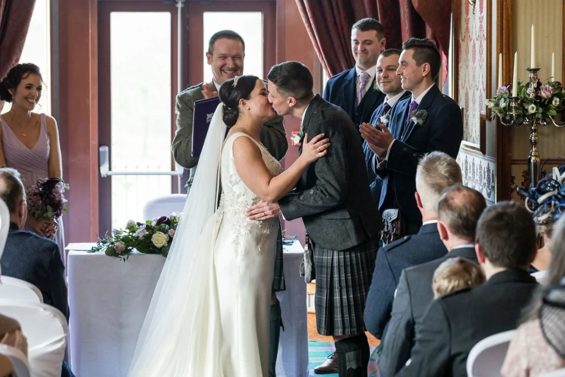 A bride and groom kiss at the altar, surrounded by smiling guests in a room with elegant decor.