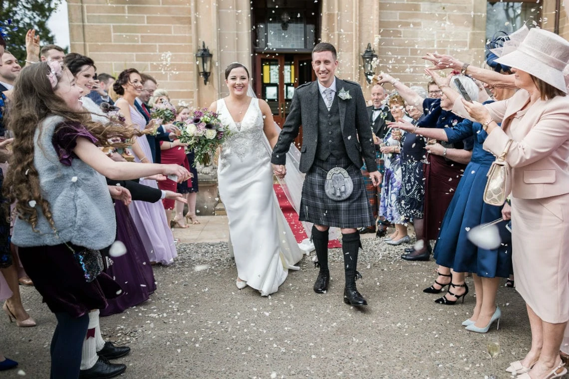 A joyful bride and groom walking through a shower of confetti, surrounded by smiling guests outside a church.