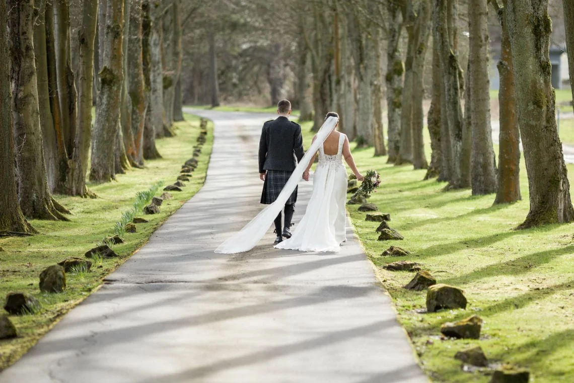 A bride and groom walking hand in hand down a tree-lined path. the bride is wearing a long white dress with a trailing veil.