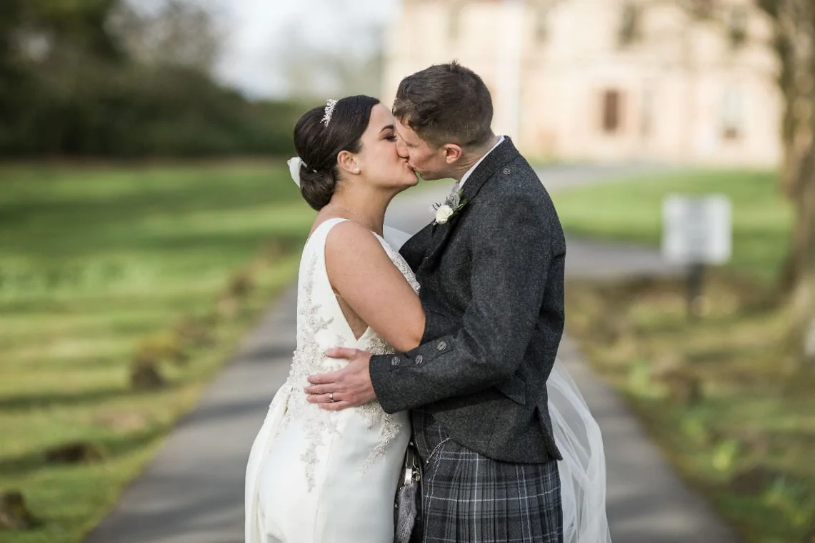 A bride in a white dress and a groom in a tartan kilt kiss tenderly on a path with a historic building in the background.