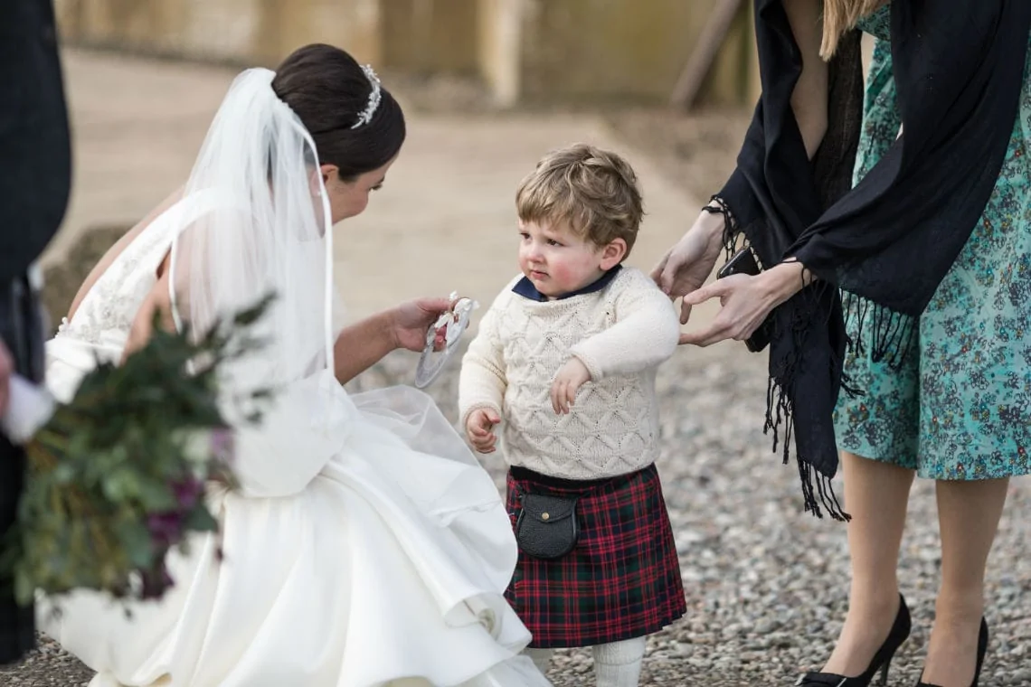 A bride kneels down to comfort a young boy in a kilt who appears upset at a wedding, while a woman assists from the side.