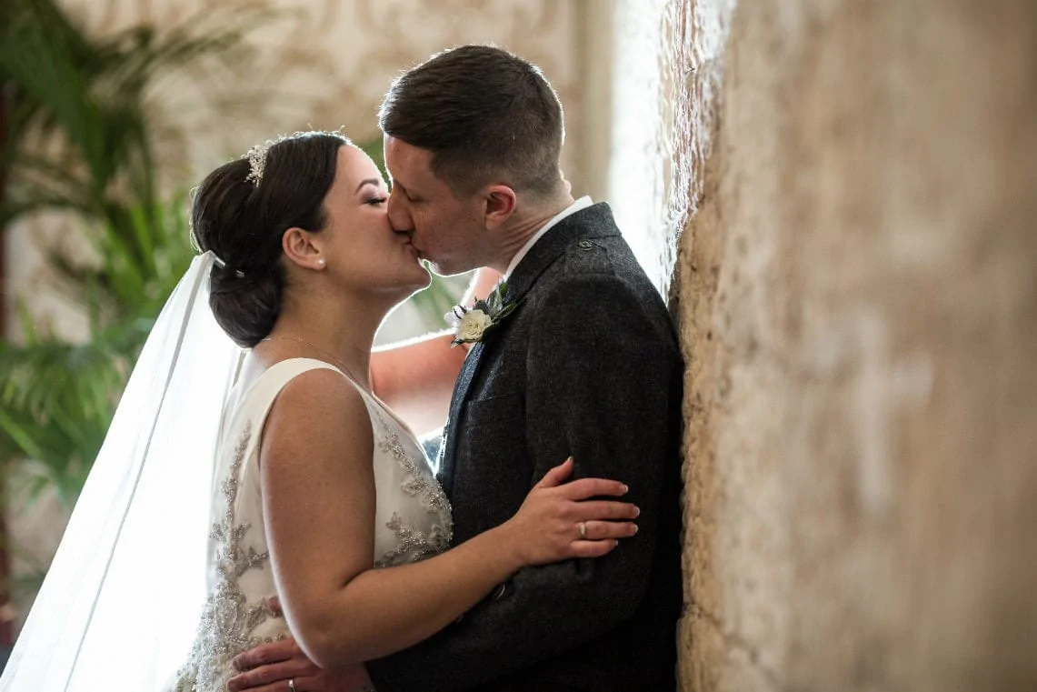 A bride and groom kissing tenderly beside a textured wall, the bride in a lace gown and the groom in a dark suit.