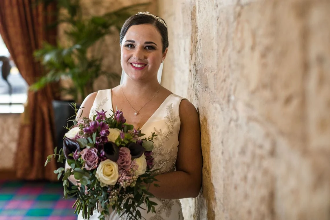 A bride in a white dress, smiling and holding a bouquet of purple and white flowers, stands beside a textured stone wall.