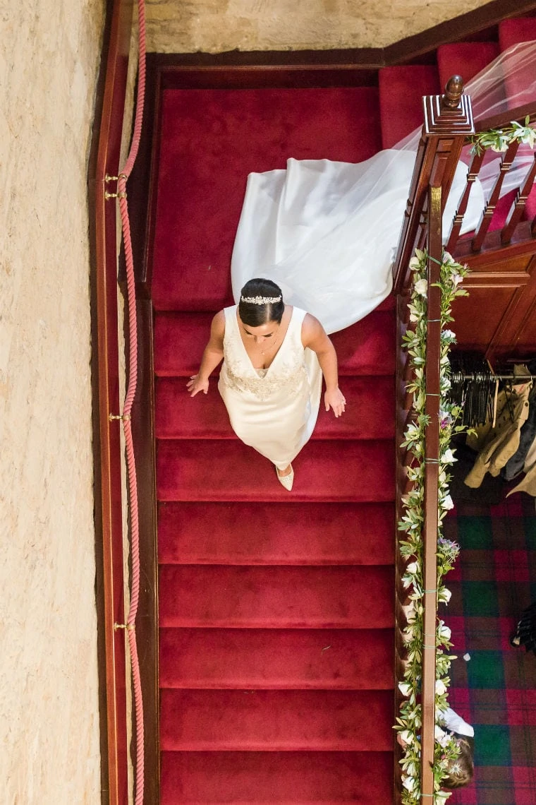 A bride walking up a red-carpeted staircase, viewed from above, with decorative plants lining the stairway.