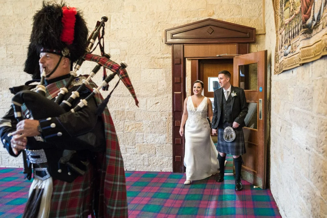 A bride and groom exit a building, smiling as they walk past a bagpiper in traditional scottish attire.