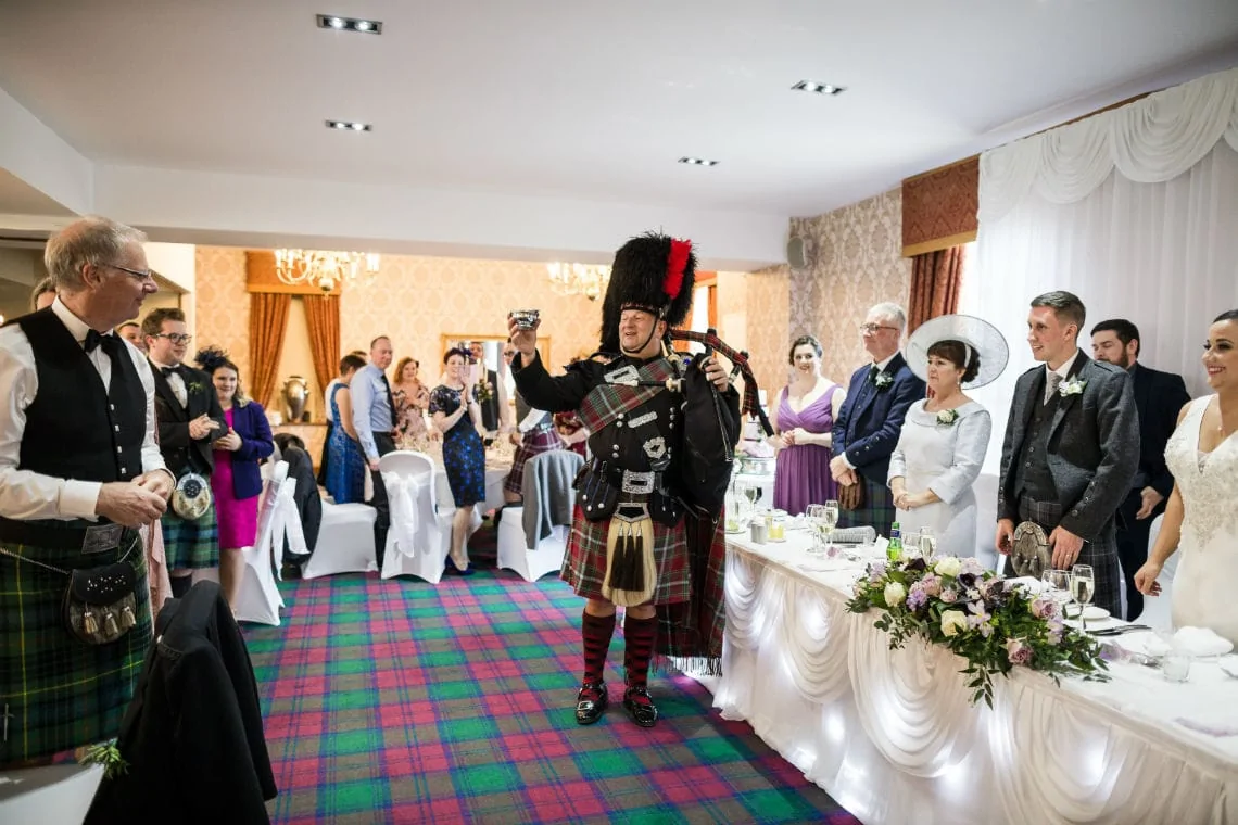 A scottish bagpiper playing in full traditional attire at a wedding reception, with guests watching in formal wear.