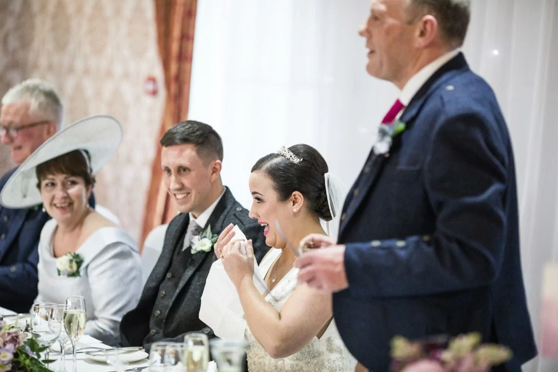 A bride joyfully laughing at a wedding reception table with groom and guests, including a man in a speech.