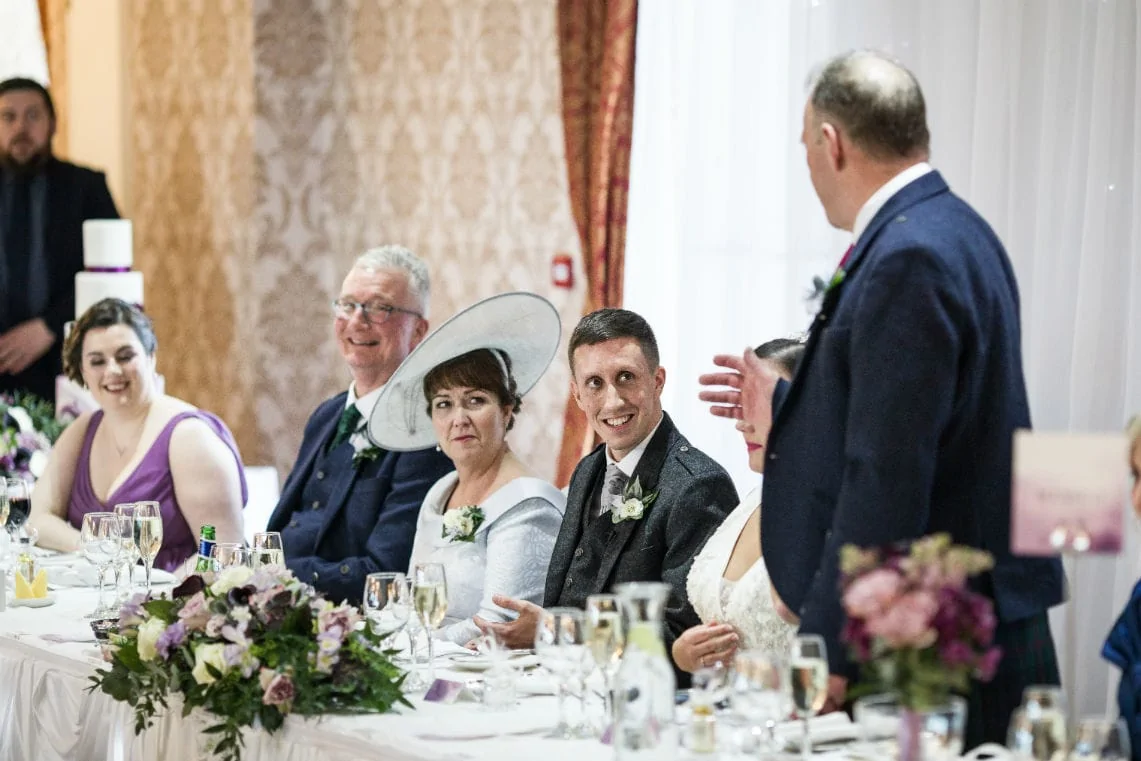 Wedding guests at a reception table smiling and listening, as a man stands speaking, with elegant floral decor visible.