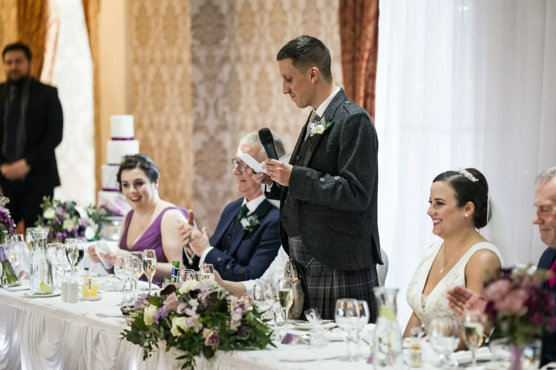 A man in a gray suit reads from a paper at a wedding table, sporting a laughter-filled audience including a man in black and smiling women in purple and white dresses.