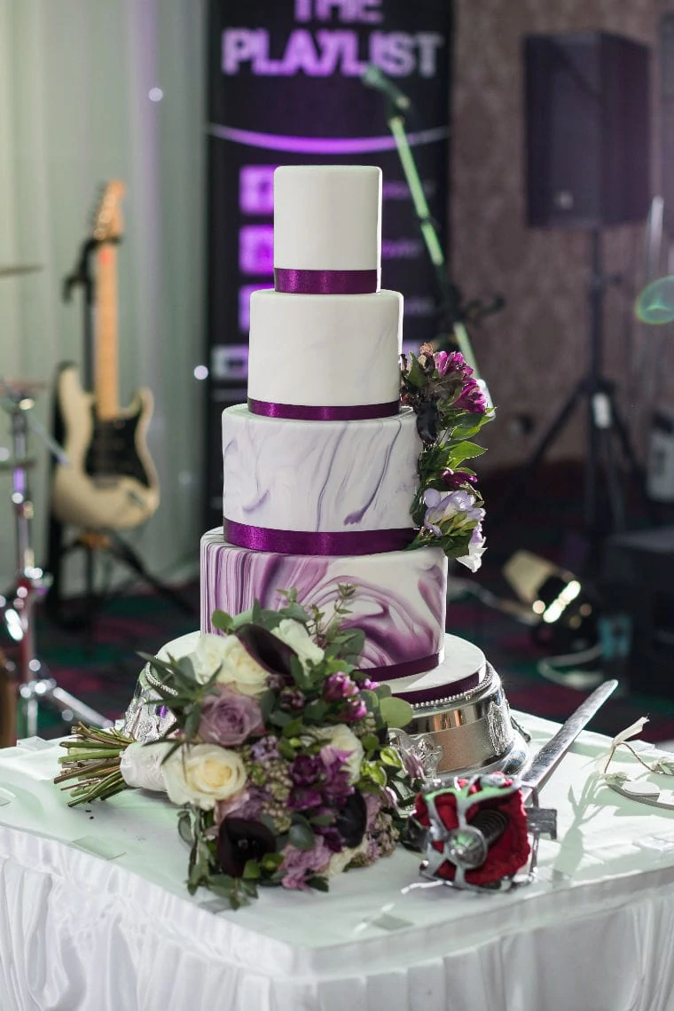 A four-tiered wedding cake with purple accents and floral decorations, displayed in front of a live band setup at a reception.