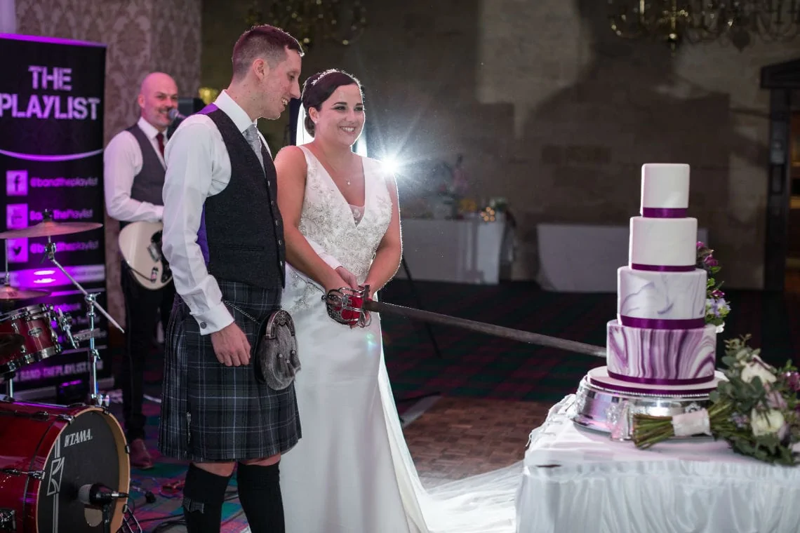 A bride and groom in wedding attire smiling and slicing a purple and white tiered cake at their reception, with a band in the background.