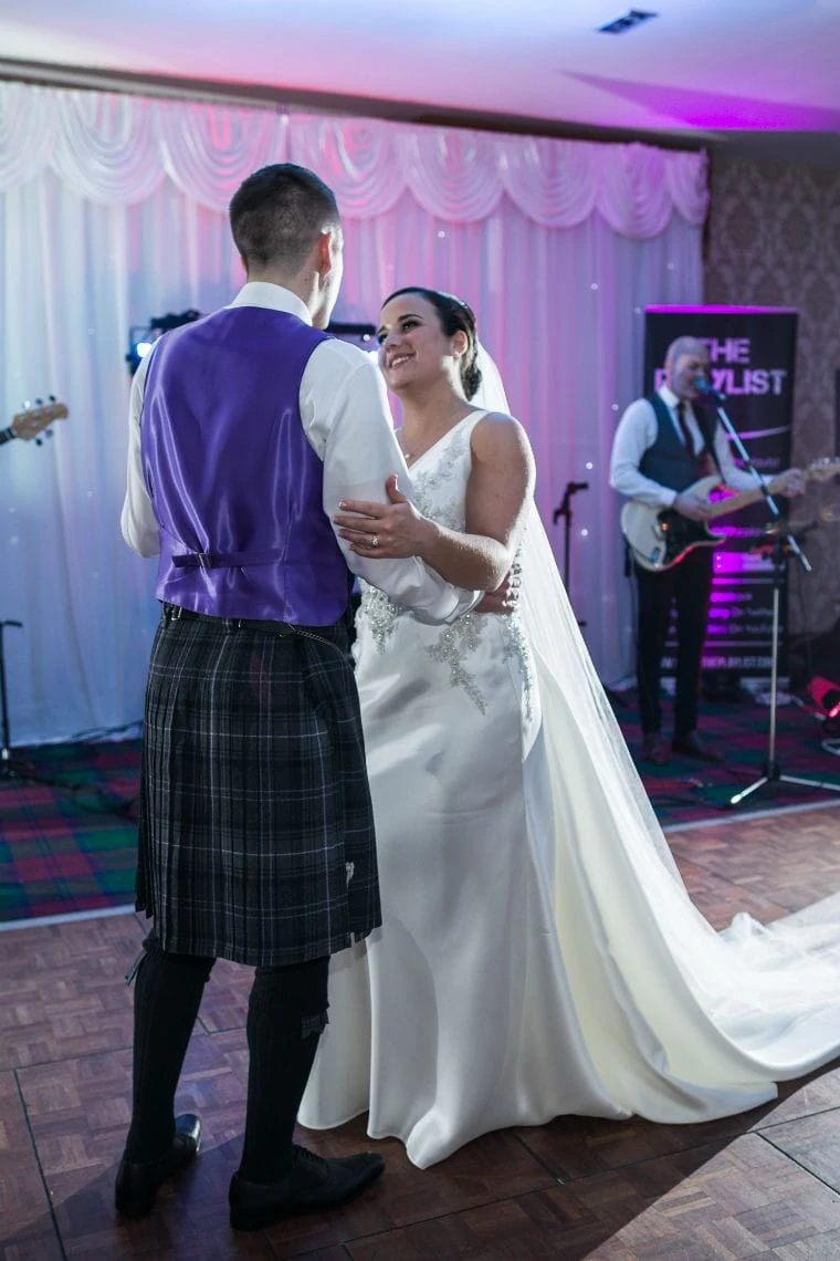 Bride and groom dancing at their wedding reception, with the groom wearing a kilt and a purple vest and the bride in a white gown, a live band performing in the background.