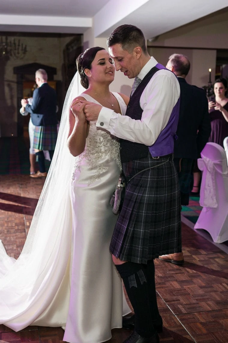 A bride and groom share a dance at their wedding, the groom in a kilt and the bride in a white gown, both smiling warmly.