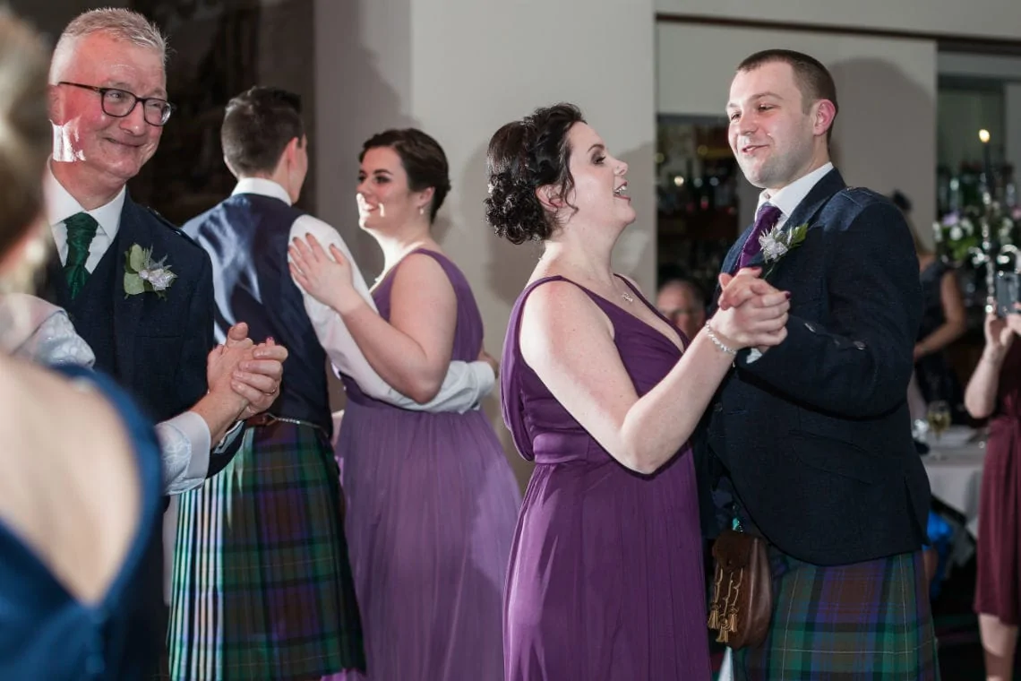 A bride and groom in traditional scottish attire dance joyfully at their wedding reception, surrounded by clapping guests.