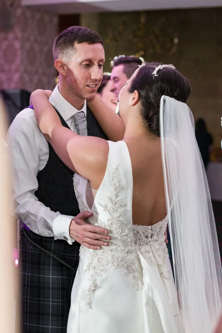 A bride and groom share a dance, the bride in a detailed white gown with a veil, and the groom in a vest and kilt.