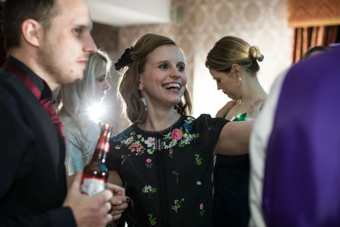 A woman in a floral dress smiling brightly at a social gathering, holding a beer, surrounded by other guests in a warmly lit room.