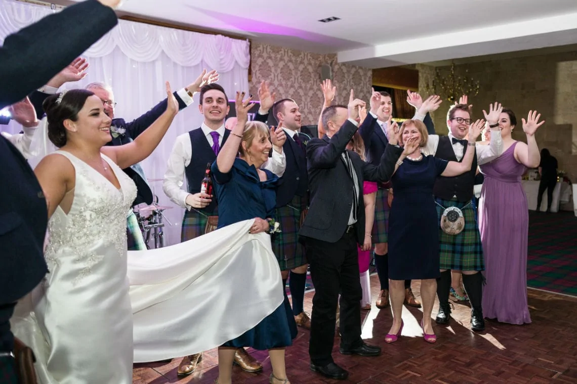 Guests joyfully dancing and waving their hands at a lively wedding reception, some wearing traditional scottish kilts.