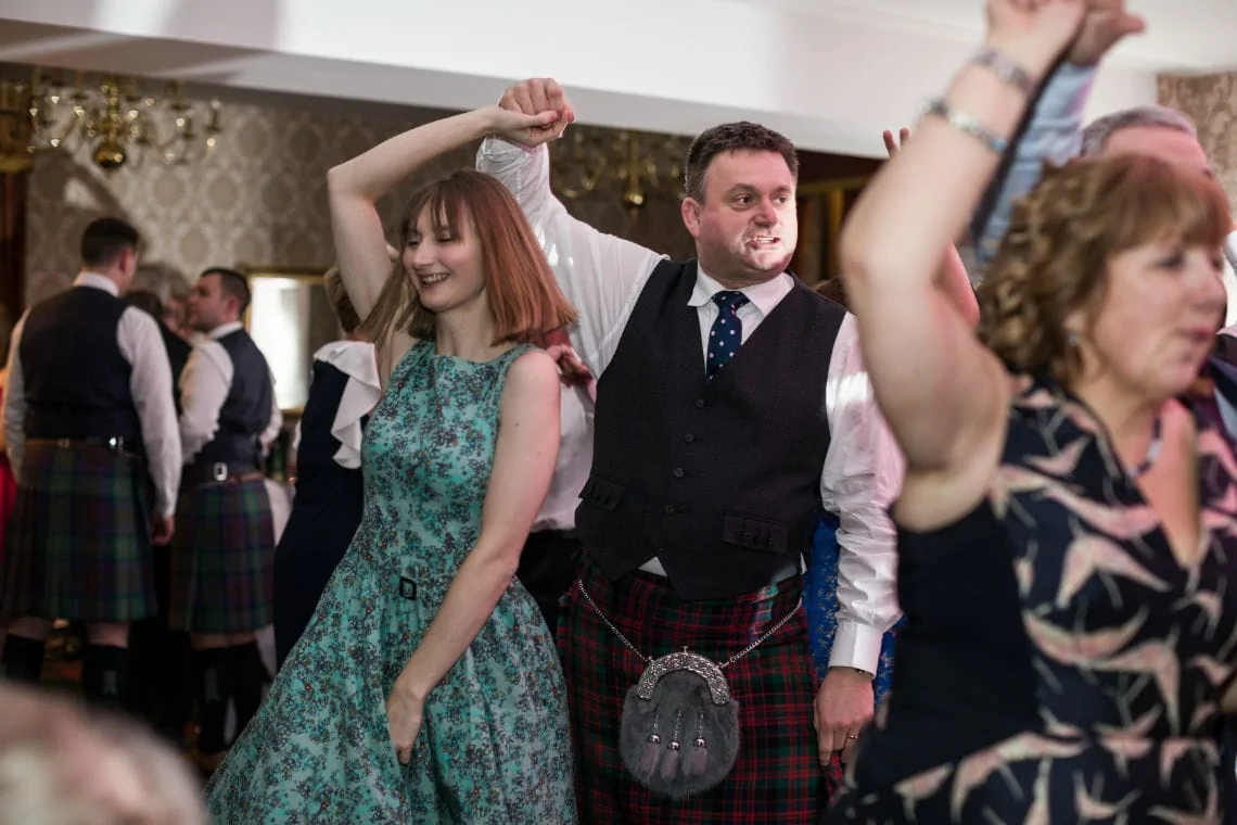 Guests in traditional scottish attire dance joyously at a wedding reception.