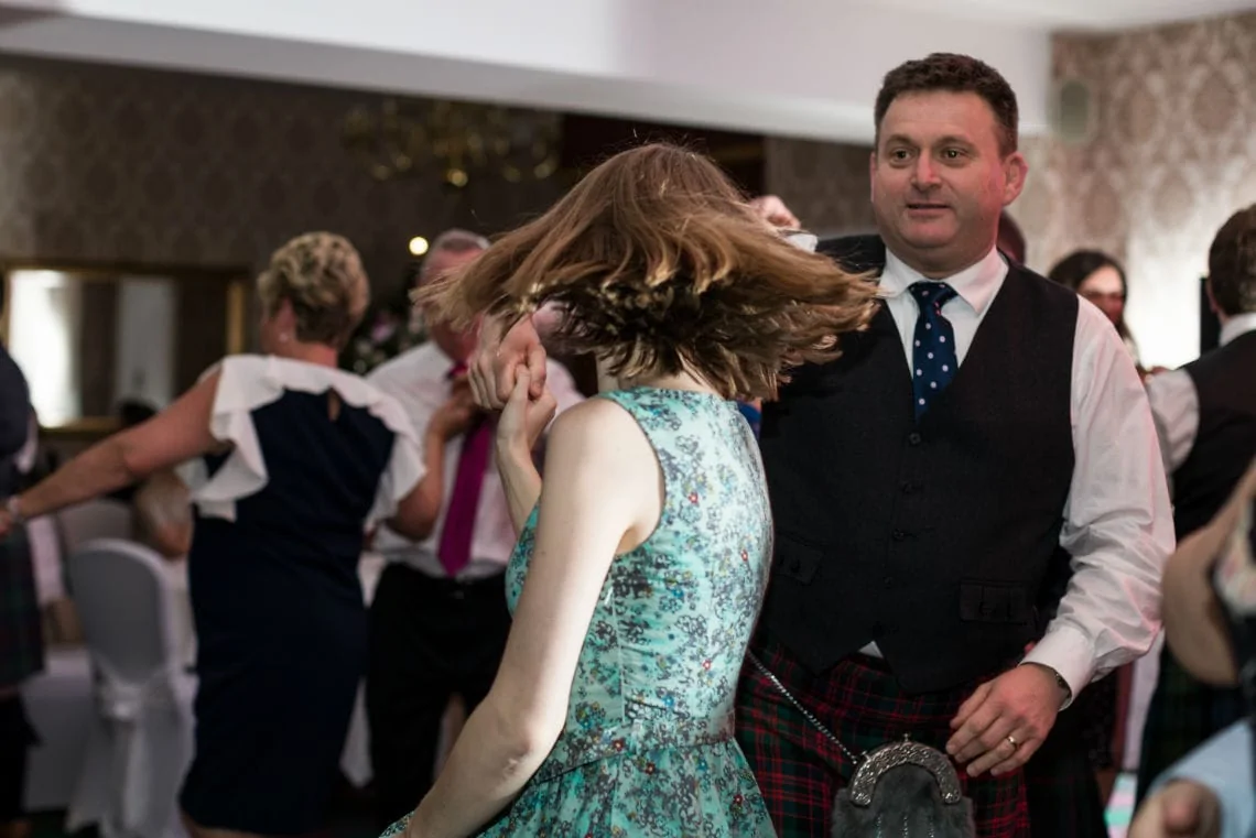 A man in a vest and kilt looks at a woman in a floral dress, twirling her hair as they dance at a lively indoor event.