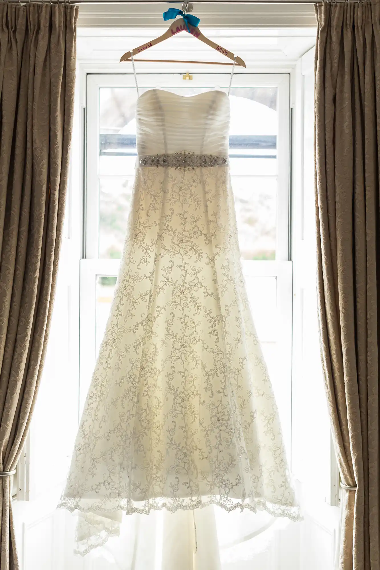 A wedding dress with lace detailing hangs on a wooden hanger in front of a window flanked by beige curtains.