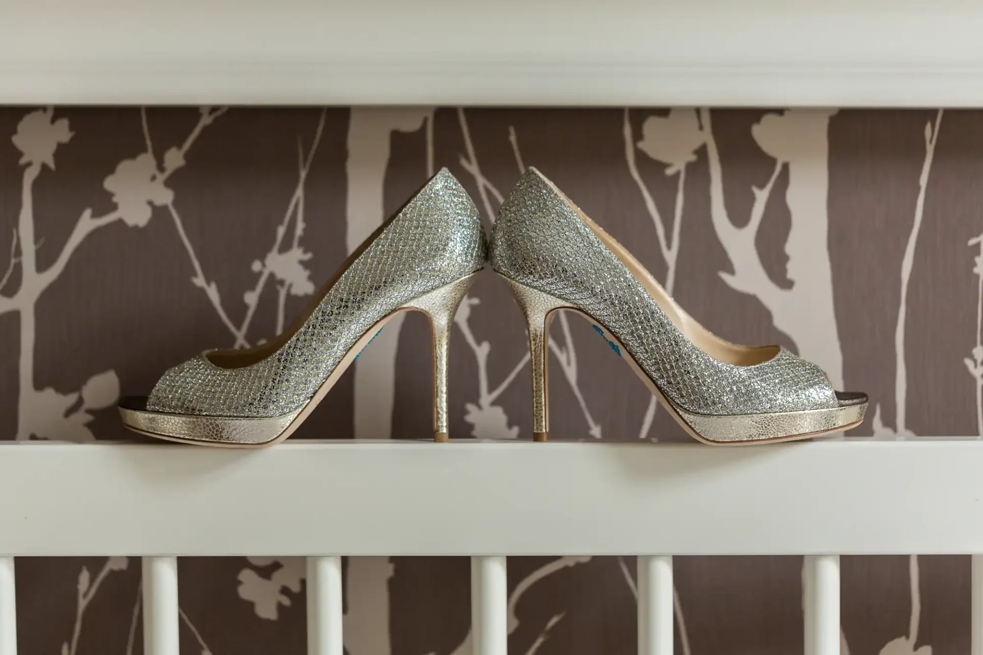 A pair of glittery silver high-heeled shoes displayed on a white railing against a patterned brown wallpaper background.