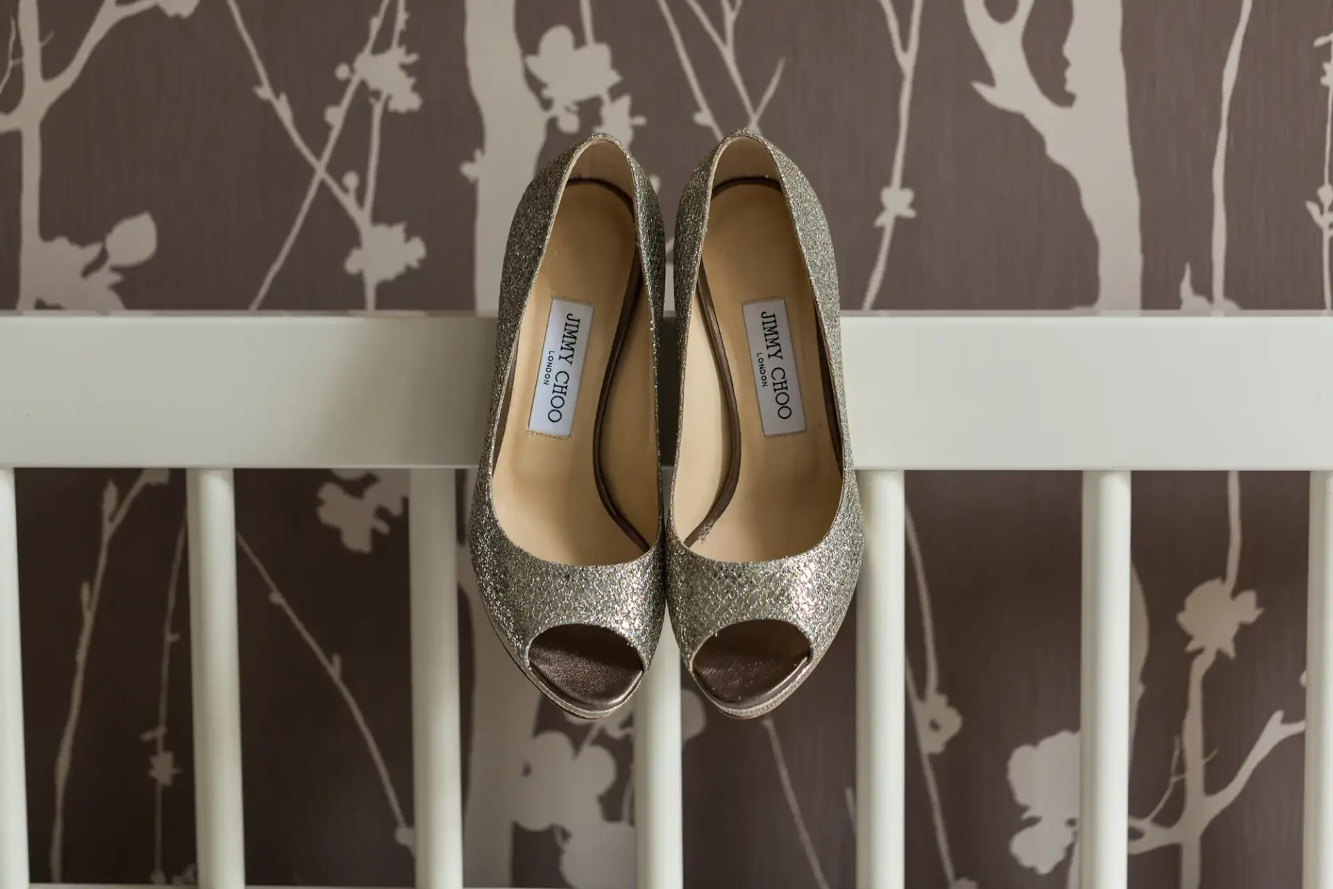 A pair of glittery jimmy choo high heels displayed on a white wooden ledge against a floral-patterned wall.