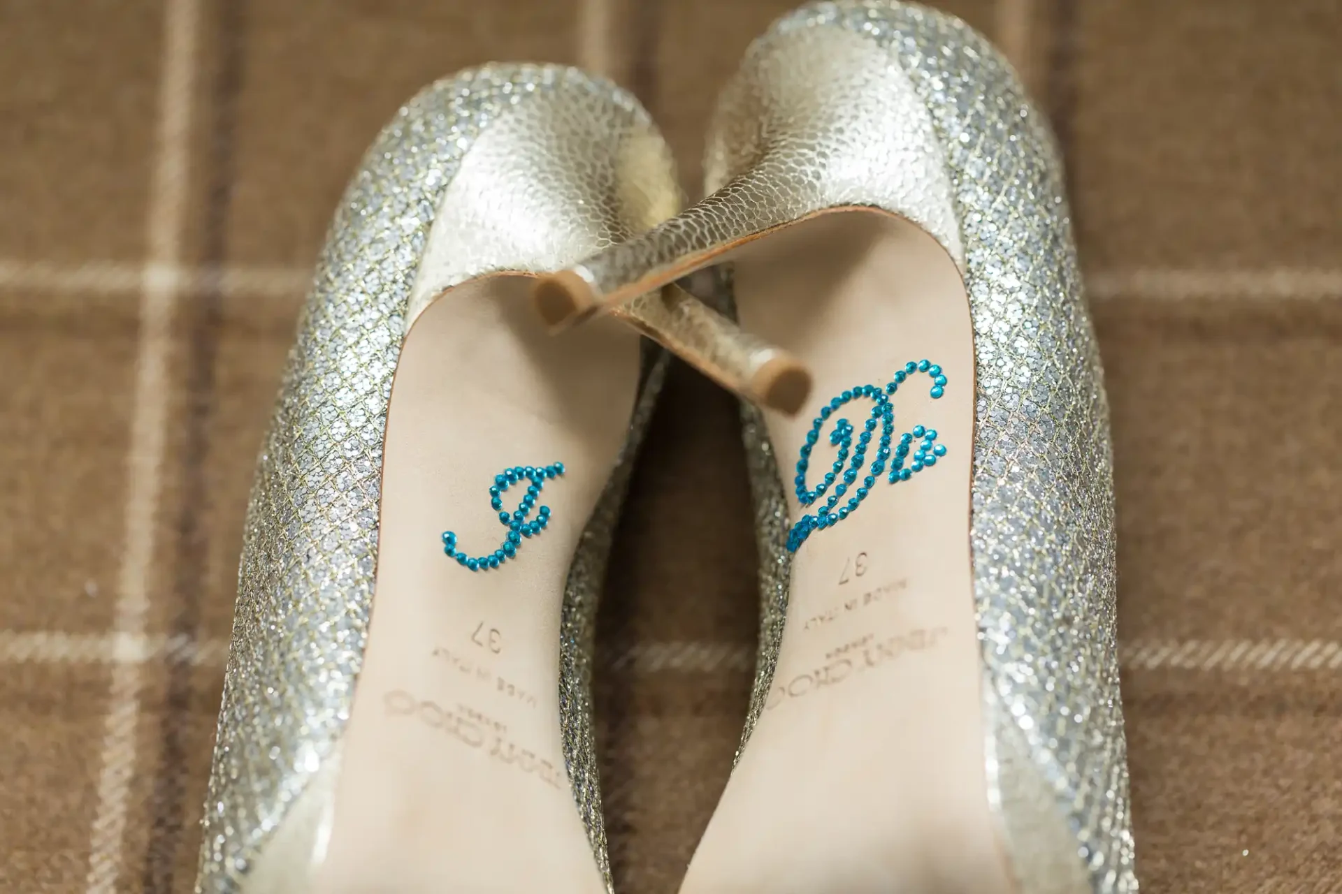 Silver glitter high-heeled shoes with "i do" written in blue on the insoles, positioned on a brown patterned background.