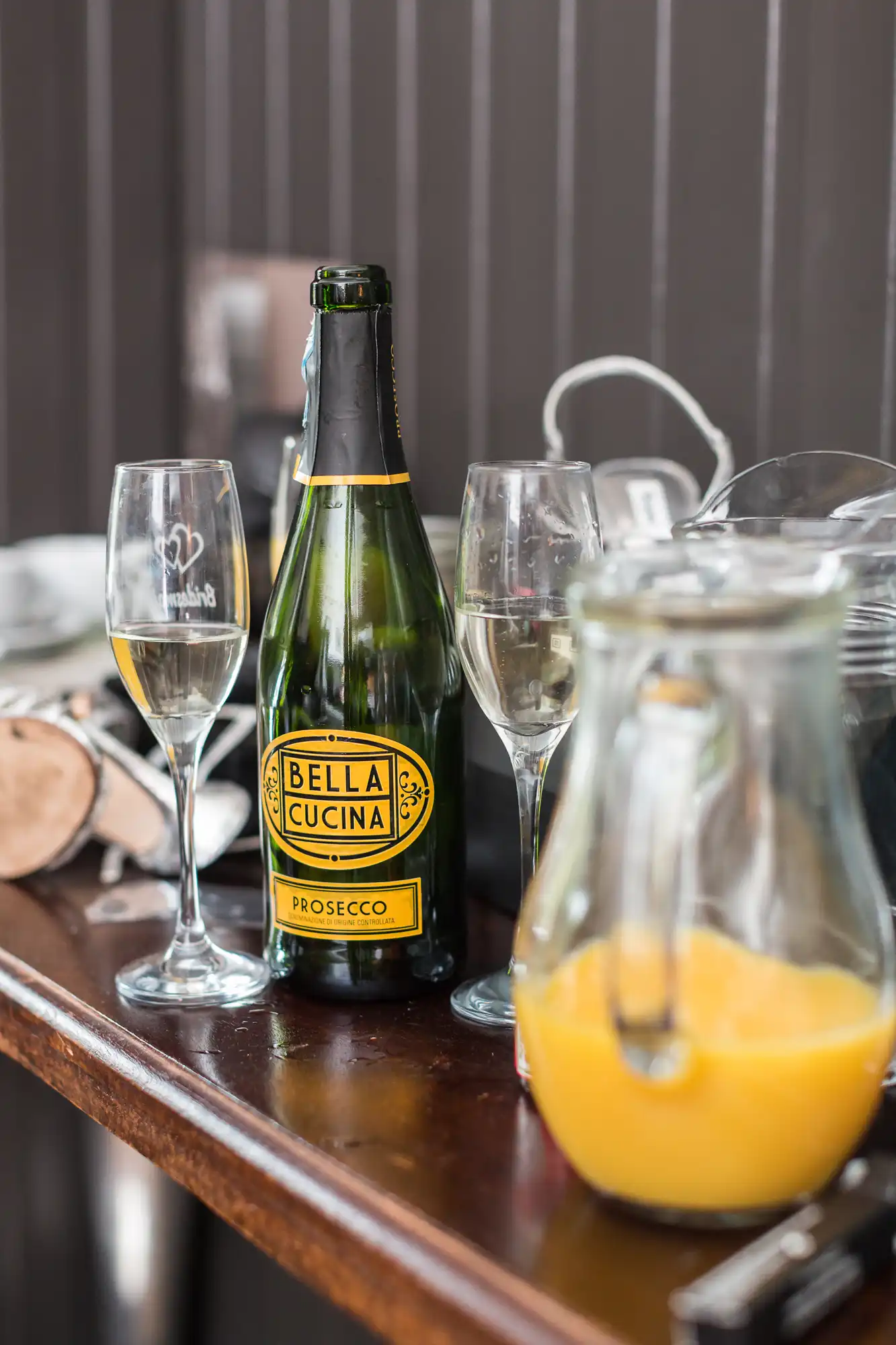 A bottle of bella cucina prosecco with two champagne glasses and a pitcher of orange juice on a wooden table.