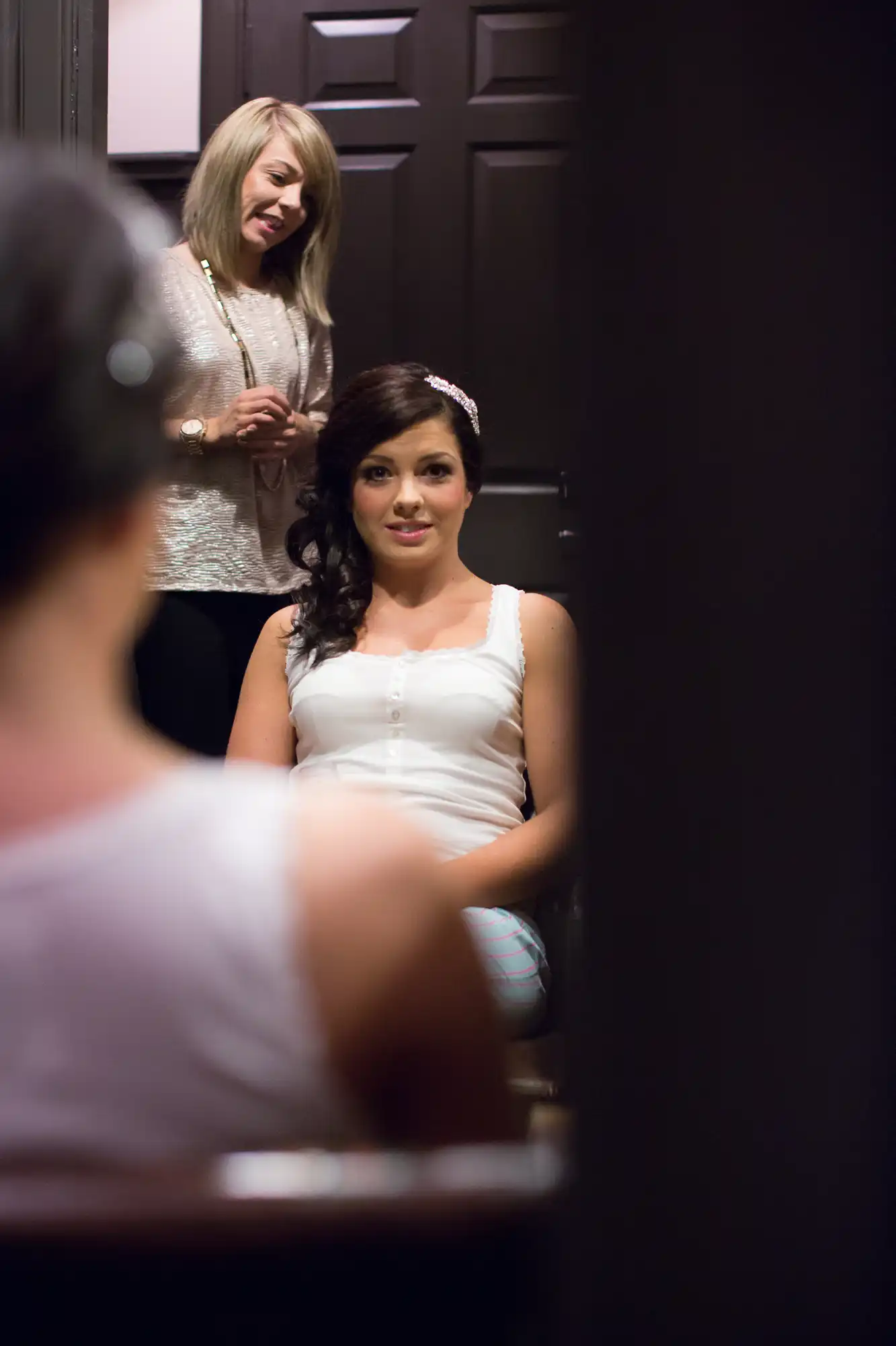 A woman sitting in front of a mirror, preparing her hair for an event, with another woman assisting her in the background.