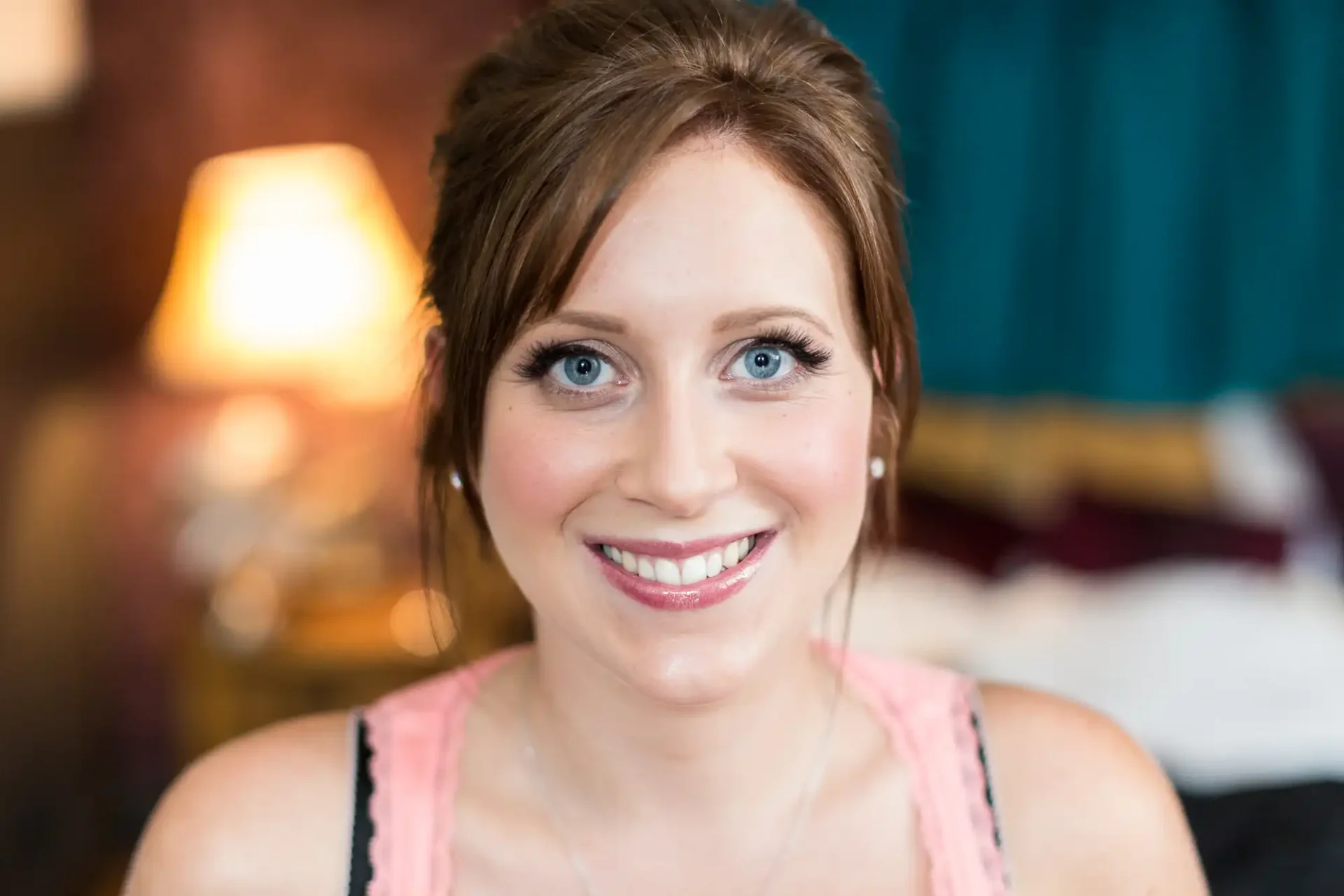 Close-up portrait of a smiling woman with blue eyes, wearing light makeup and a pink top, indoors with a softly blurred background.