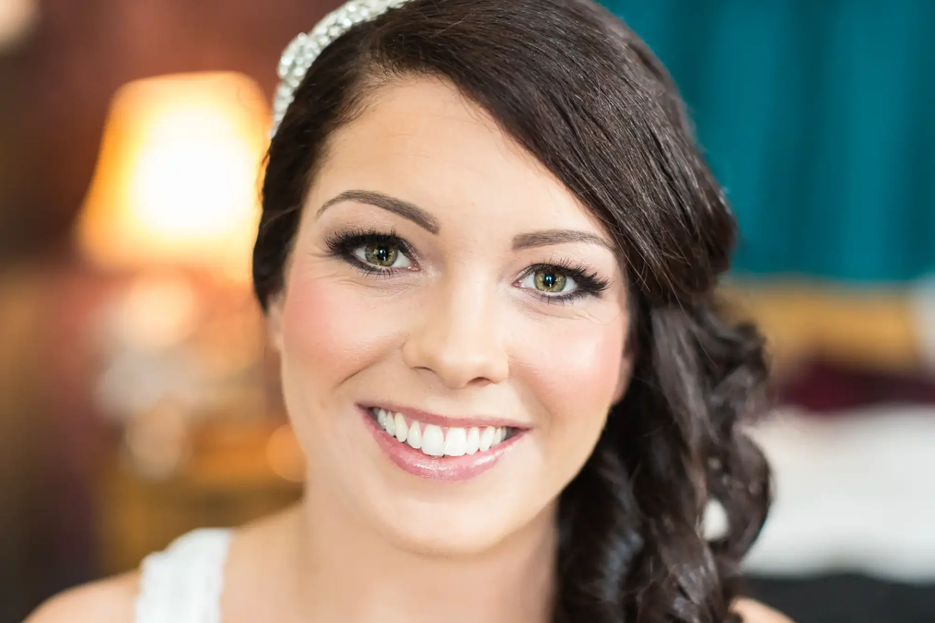 Close-up of a smiling bride with dark hair, wearing a tiara, elegant makeup, and earrings, in a warmly lit room.