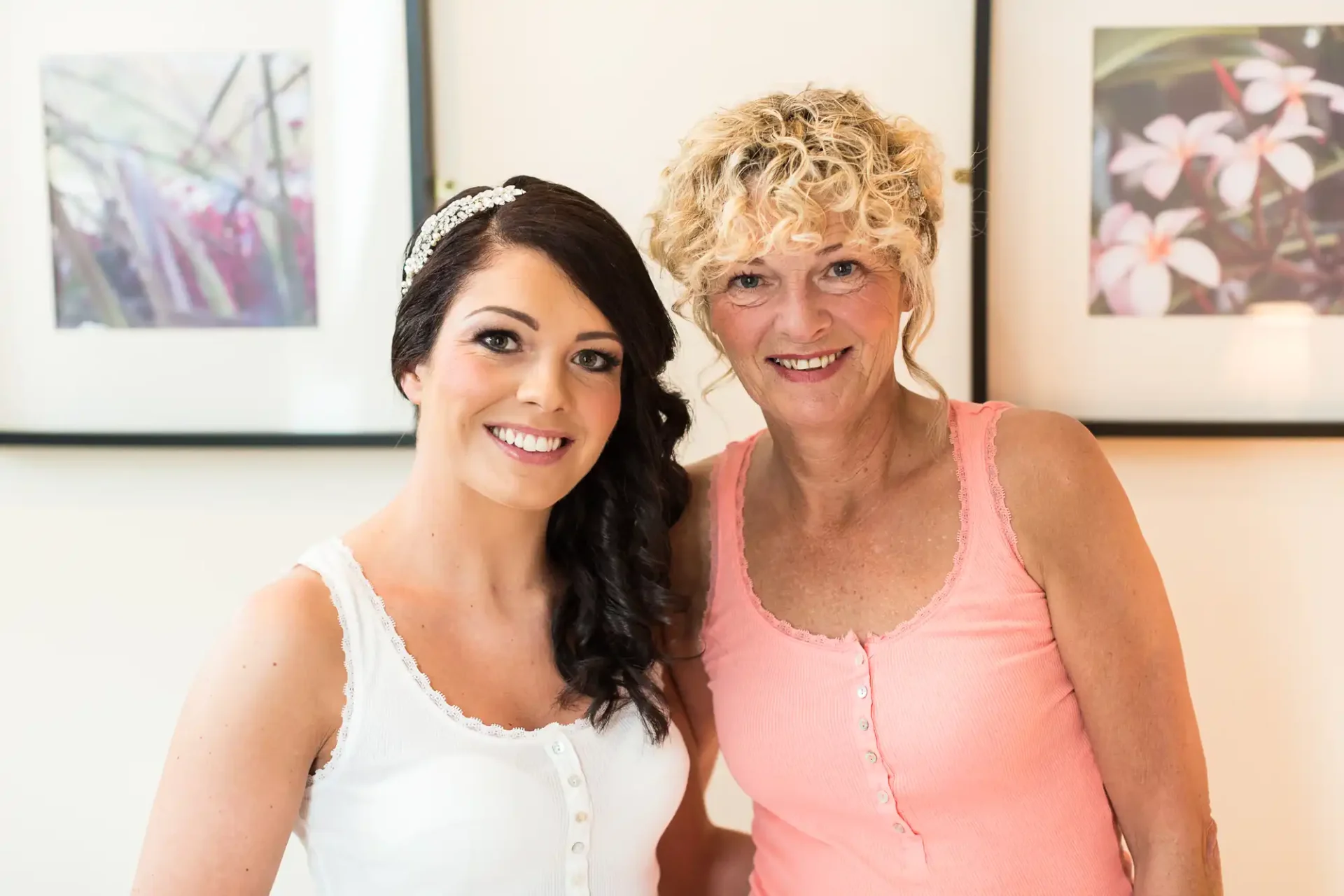 Two women, one younger with dark hair and a tiara, and an older with curly blonde hair, smiling together indoors near floral artwork.