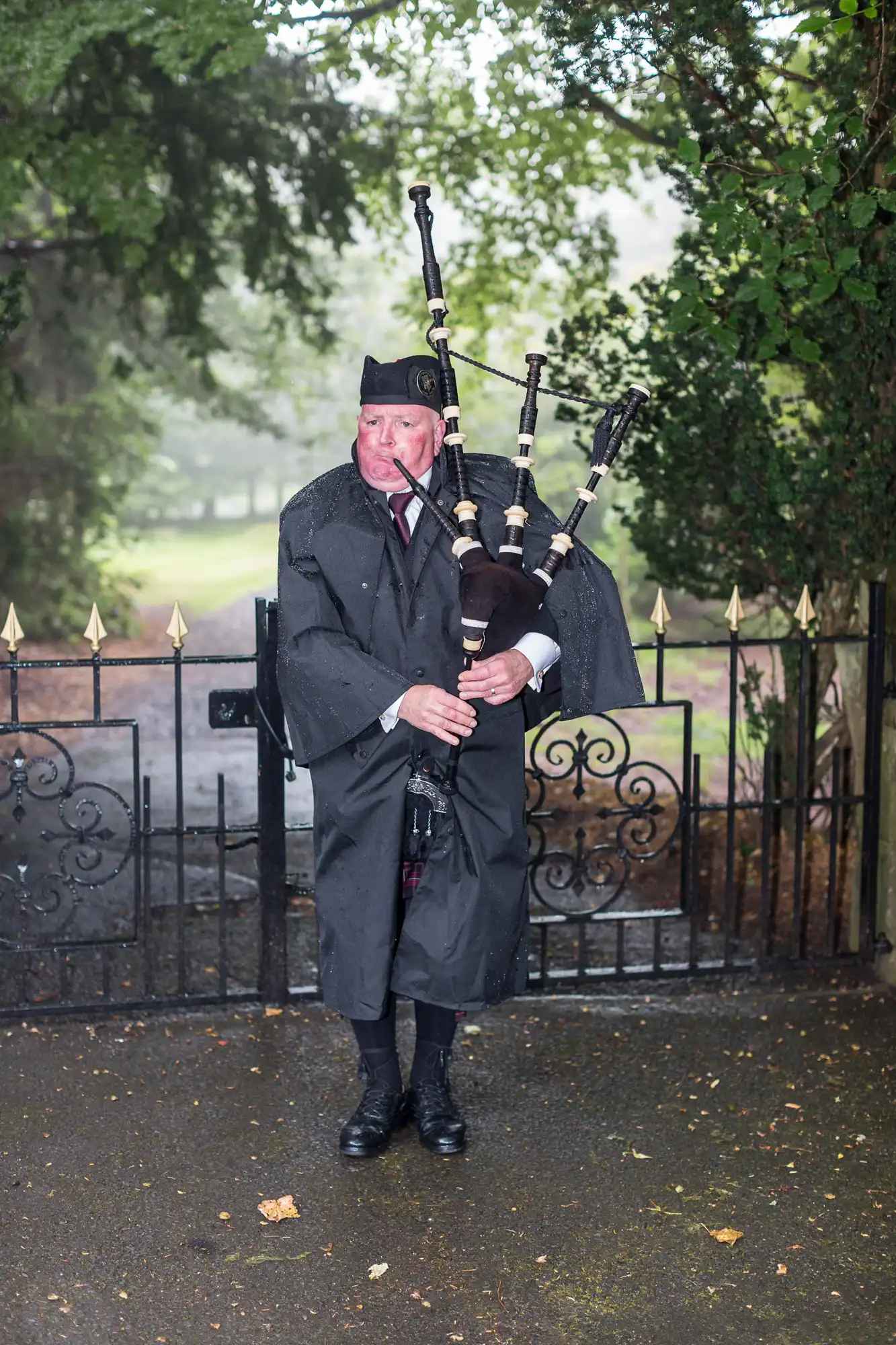 An elderly man in traditional scottish attire playing bagpipes in the rain near a gate.