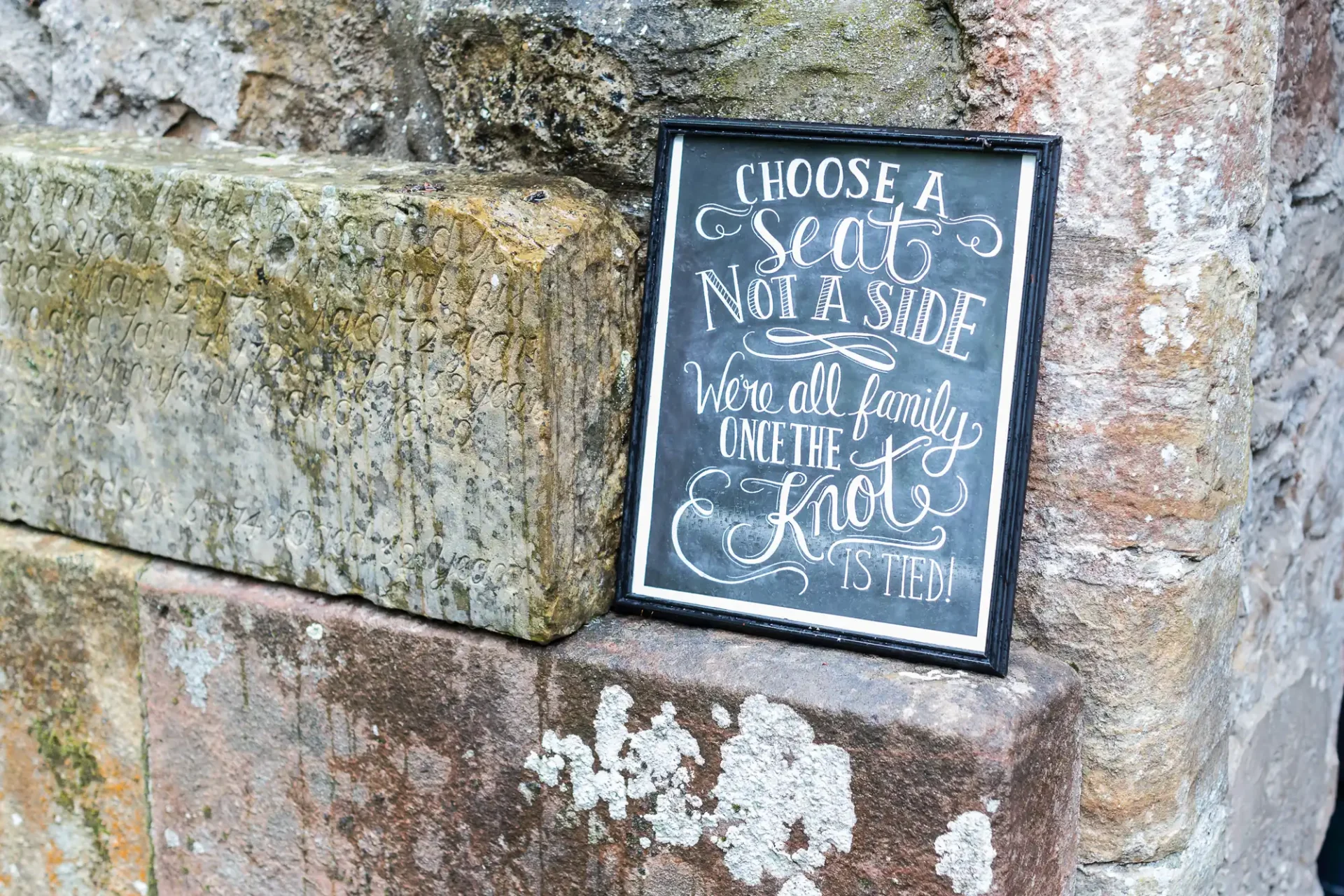 A framed sign on a weathered stone wall reading "choose a seat not a side, we're all family once the knot is tied!.