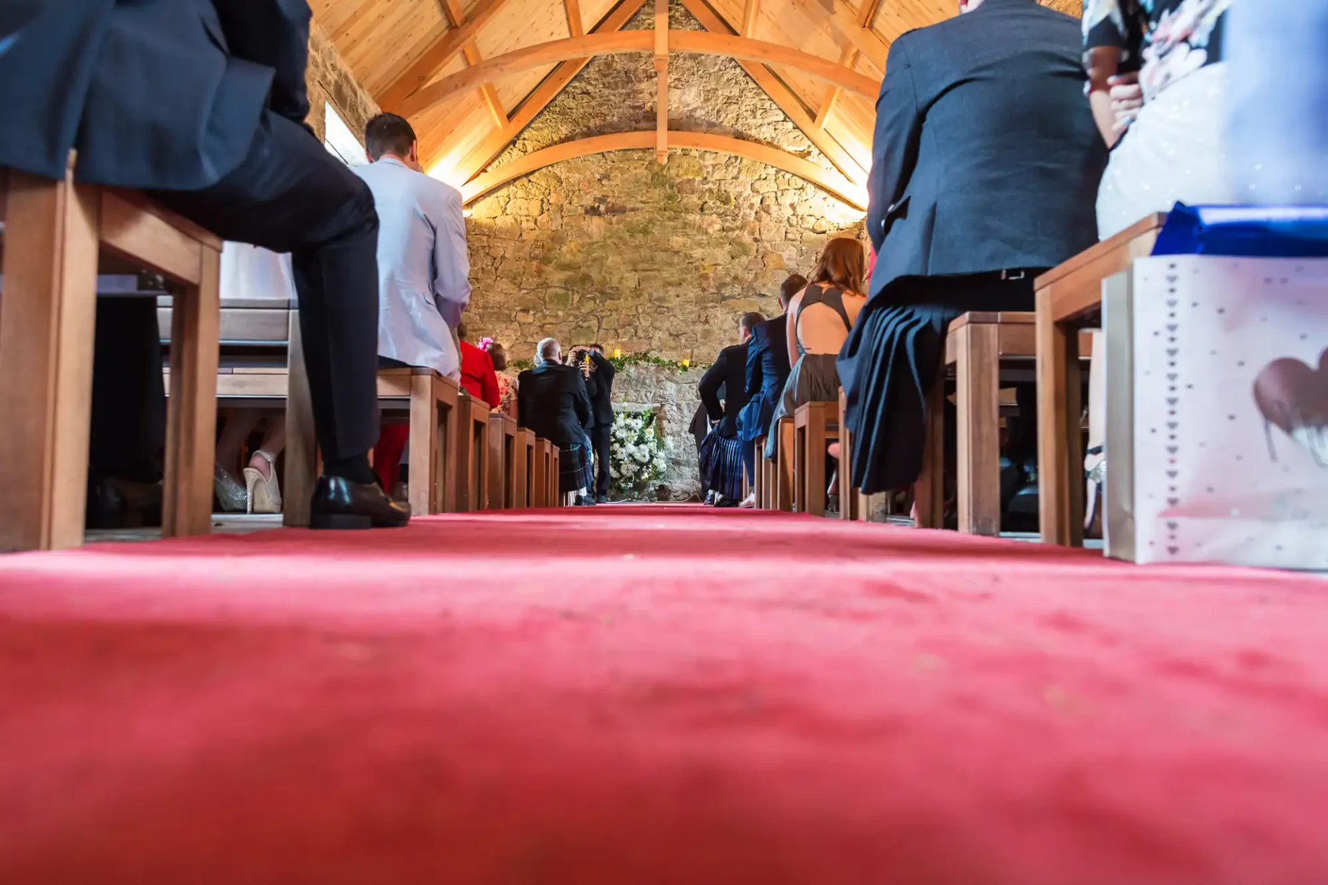 Guests line up inside a stone chapel with a red carpet leading to the altar during a wedding ceremony.