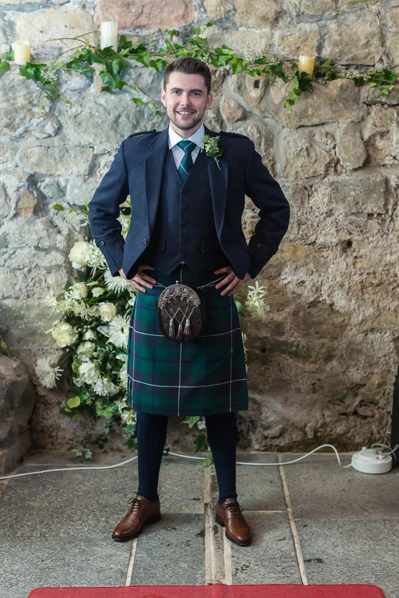 A man in a traditional scottish kilt and jacket standing with a smile in a rustic stone-walled room decorated with candles and flowers.