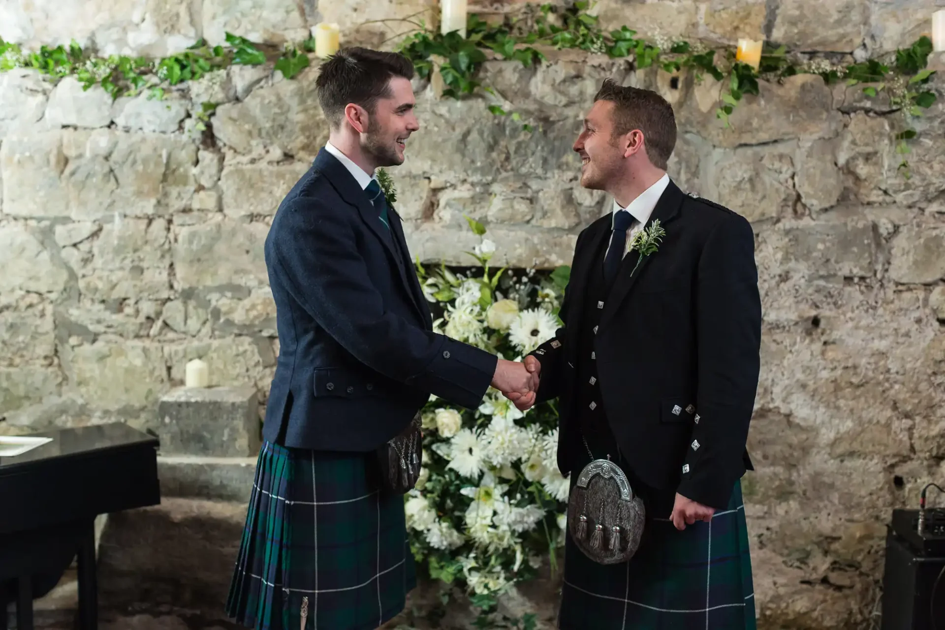 Two men in traditional scottish kilts shaking hands at a wedding ceremony, standing in front of a stone wall adorned with ivy.