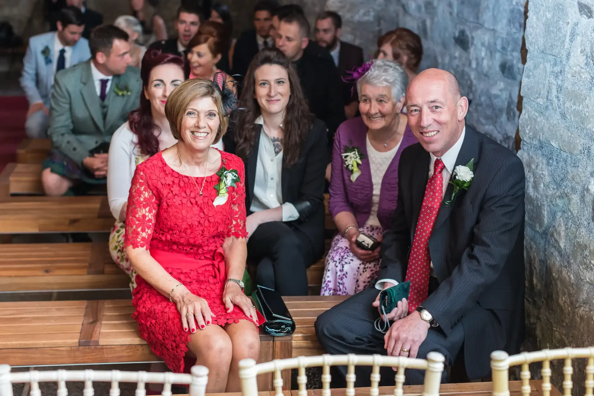 Group of guests sitting in a church pew at a wedding, smiling towards the camera, including an older couple in the front row with flowers on their outfits.