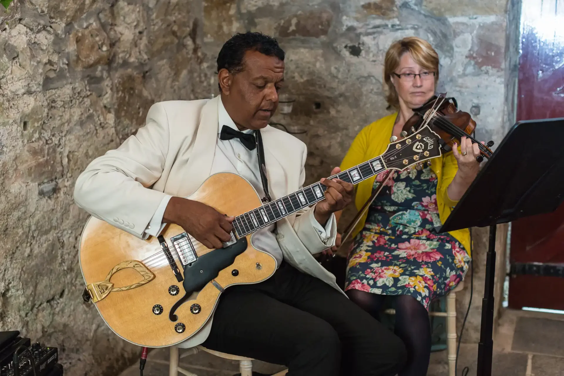 A man in a white suit plays an electric guitar while a woman in a floral dress plays a violin in a rustic setting.