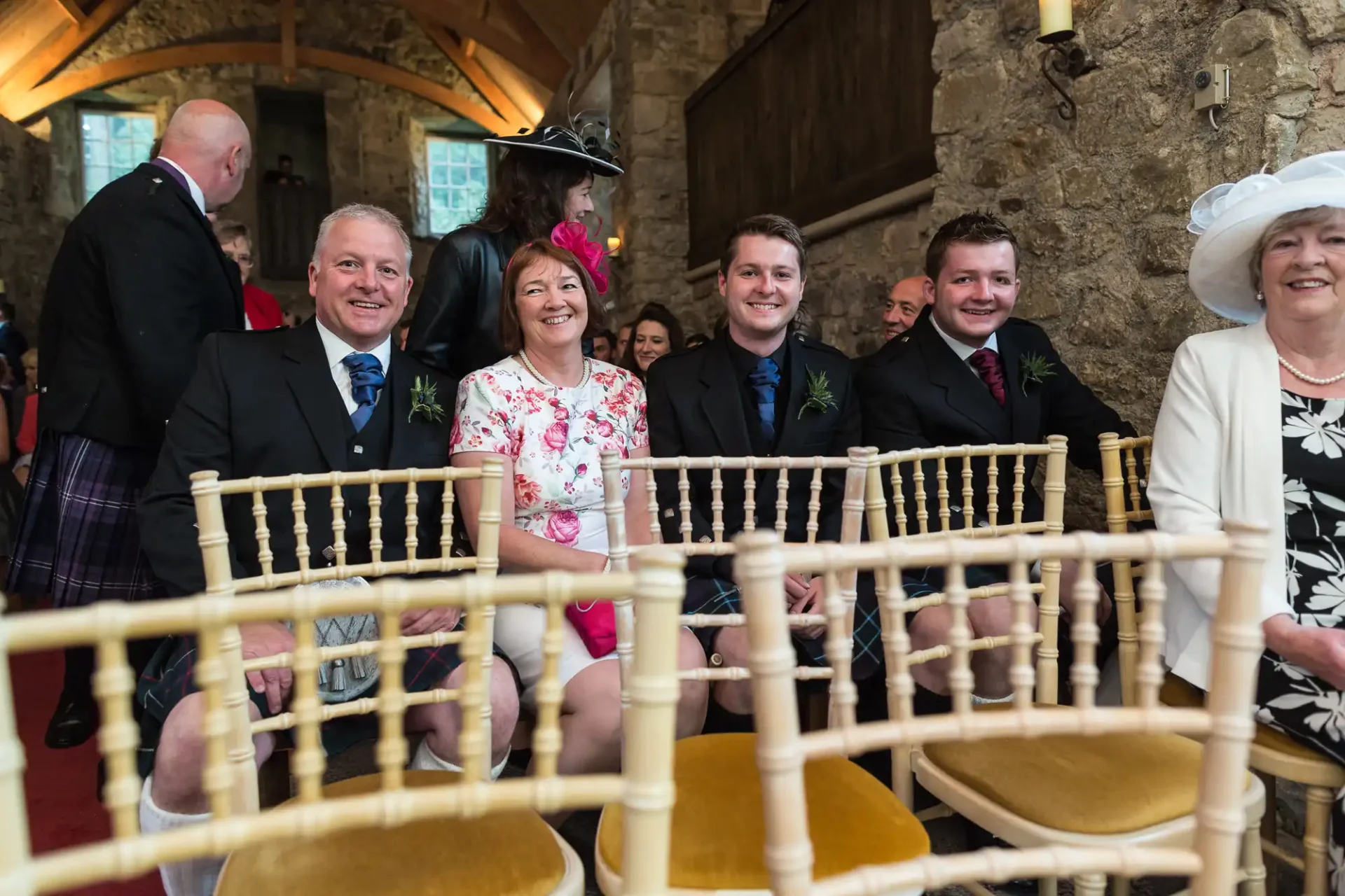 Five guests smiling and seated in a row at a wedding, with other attendees in the background, in a historic venue with stone walls.