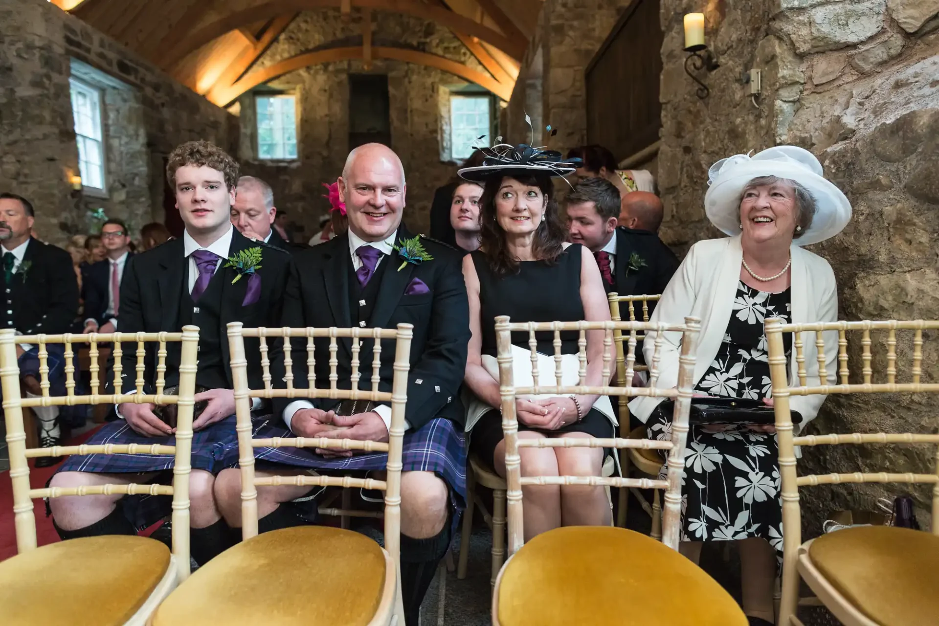 Guests at a wedding ceremony in a rustic stone hall, featuring a young man in a tartan kilt and others in formal attire, smiling as they await the event.