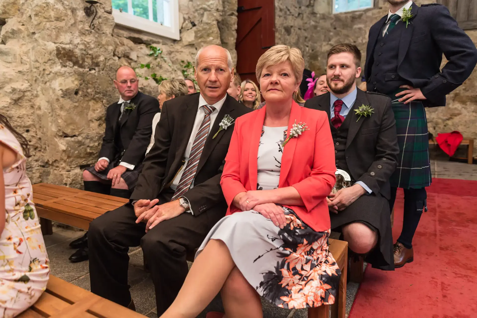 A mature couple sitting on a bench in a rustic stone church, the man in a suit and the woman in a coral blazer and floral skirt, both looking back towards the camera.