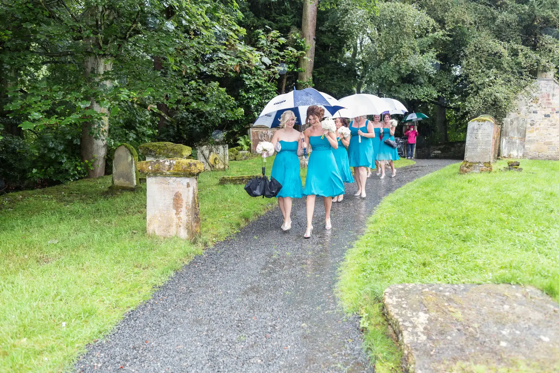 Women in turquoise dresses walk along a cemetery path with umbrellas, smiling and chatting.