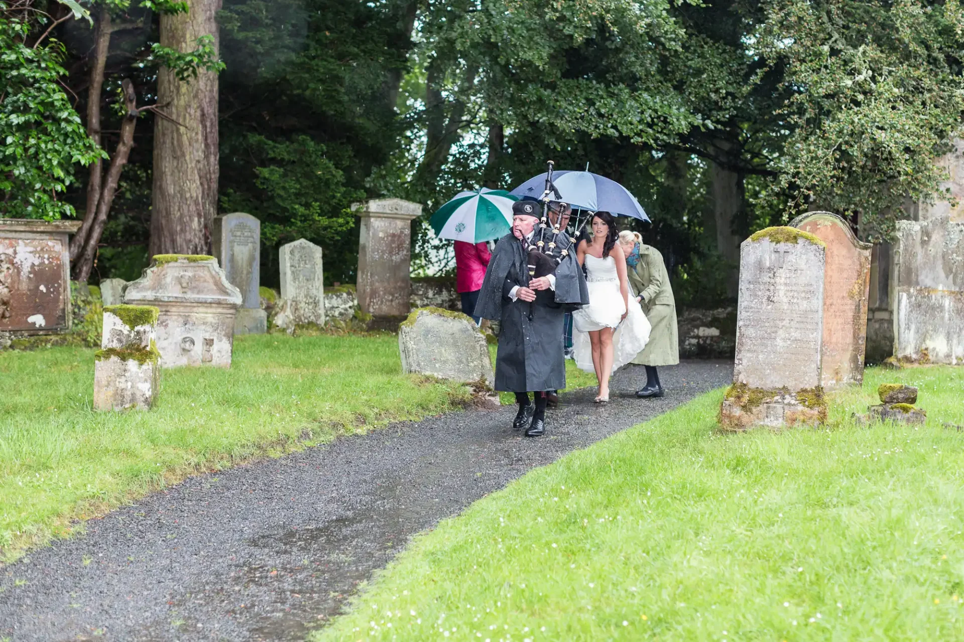 People walking on a cemetery path with umbrellas on a rainy day, dressed for a formal event amidst old tombstones.