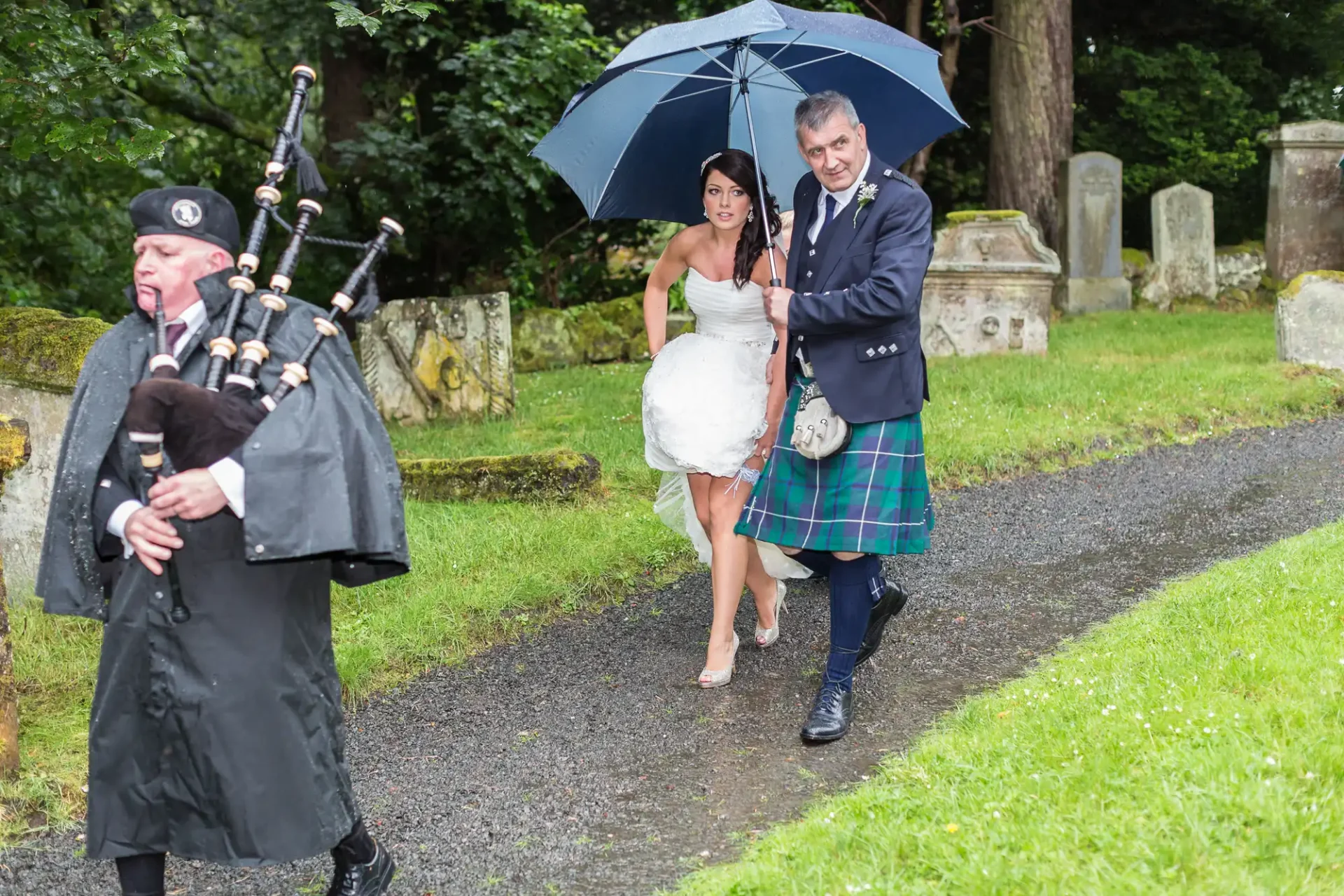 A bride and groom, the man in a kilt, walk under an umbrella led by a bagpiper on a rainy day.