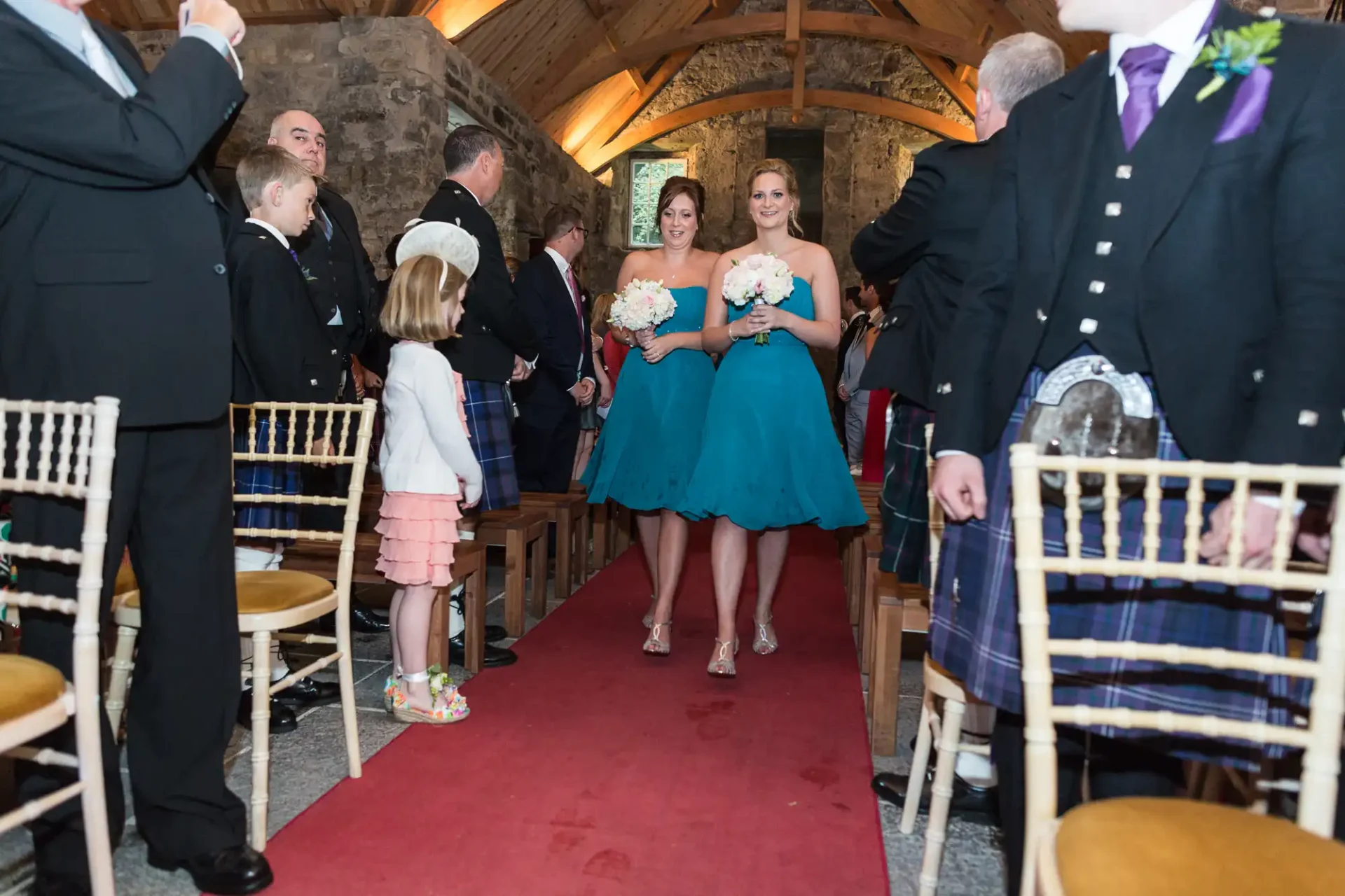 A wedding procession inside a stone chapel, with guests in formal attire, including kilts, watching two bridesmaids in blue dresses leading the way.
