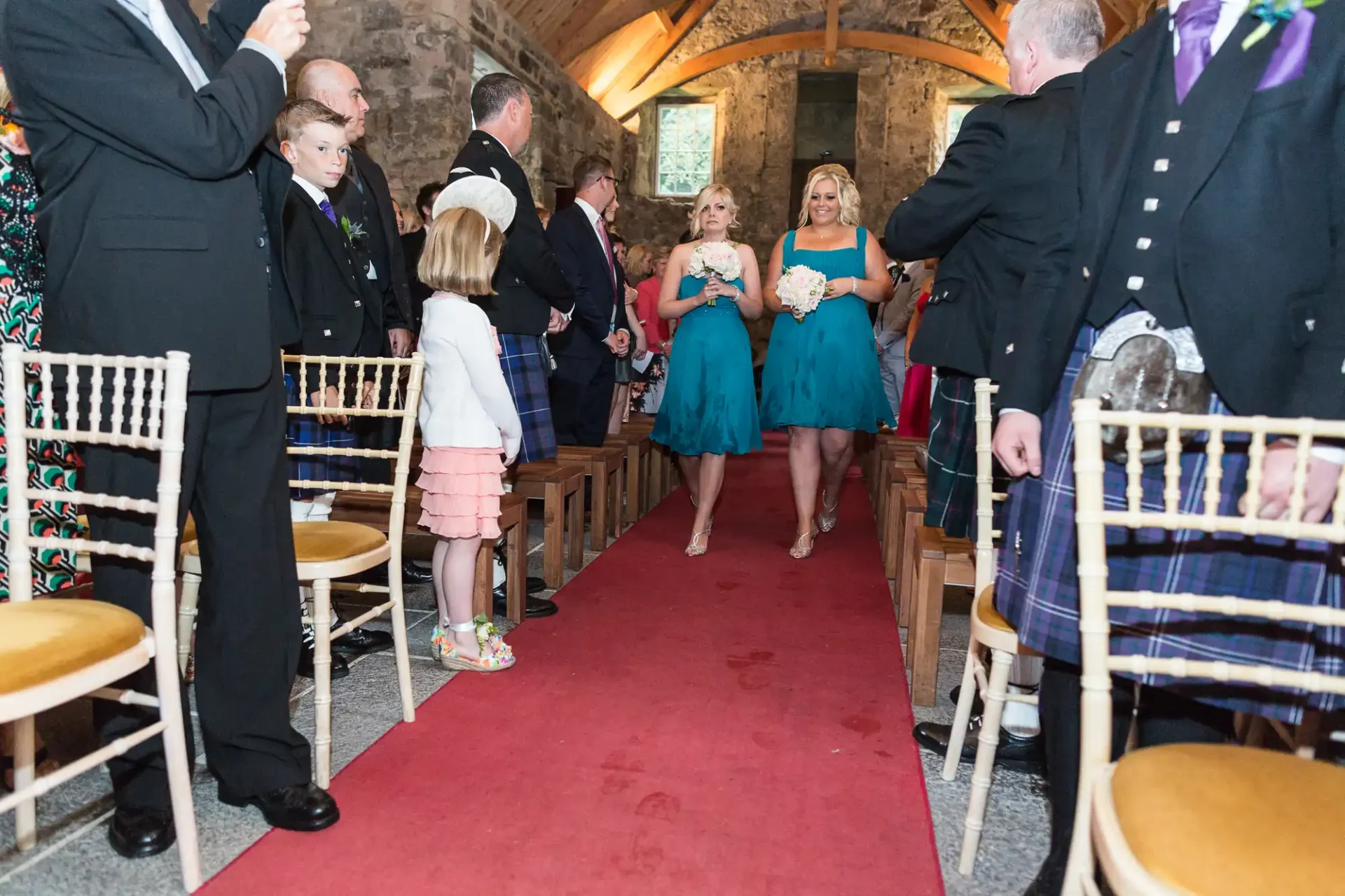 Guests in formal attire, including kilts, stand inside a stone chapel as a woman in a blue dress walks down a red carpeted aisle.